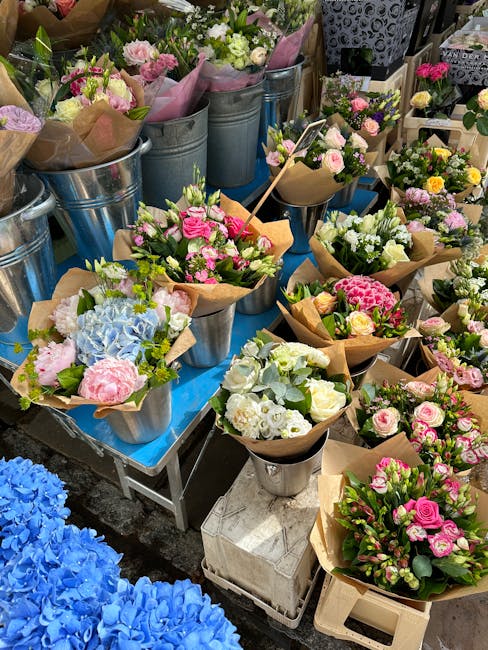 A vibrant display of fresh floral arrangements arranged in metal buckets, glass vases, and paper-wrapped bouquets on a blue-covered table. The flowers include pastel pink roses, white and pale green f